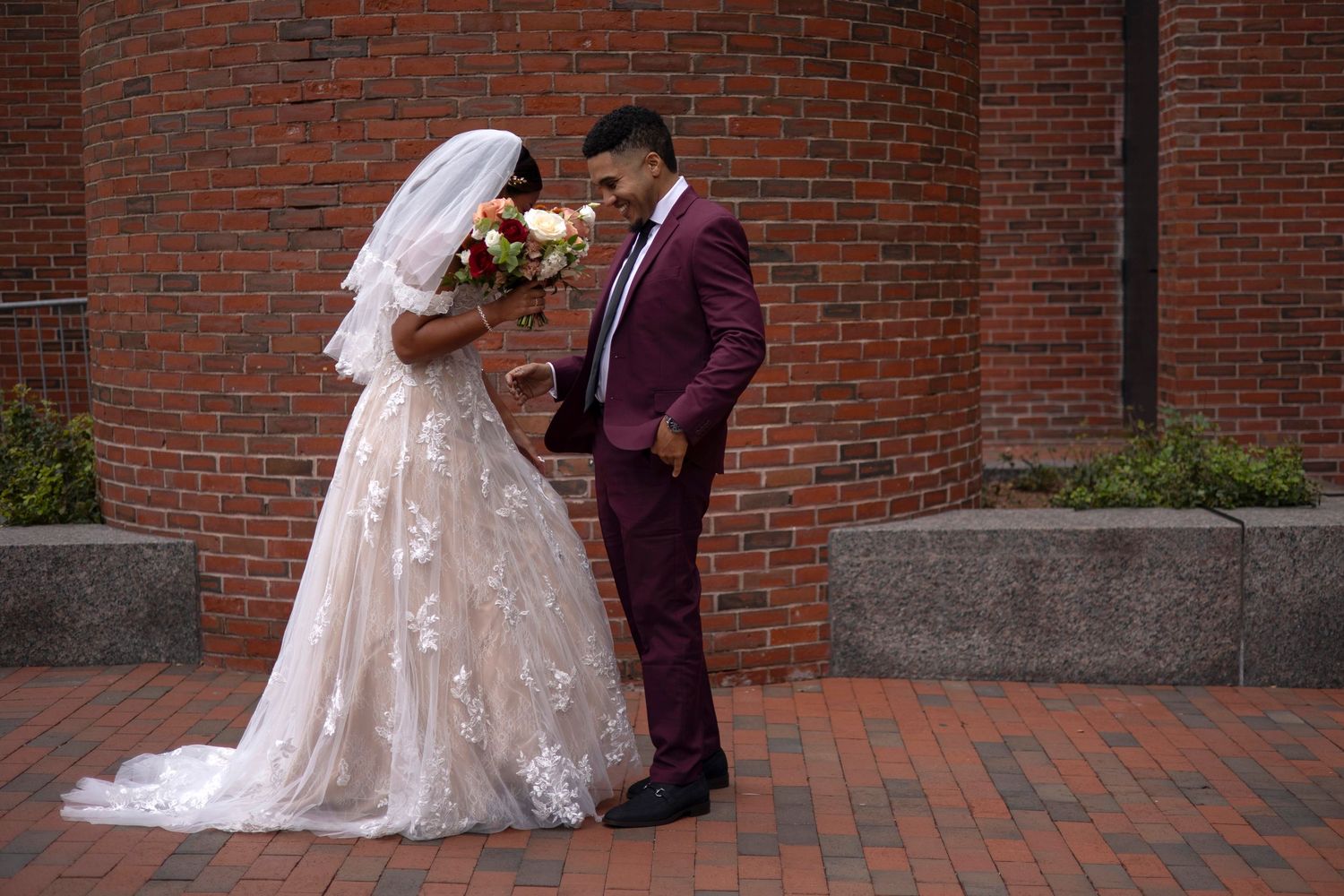 A romantic wedding couple poses against a rustic brick wall backdrop in an outdoor setting.
