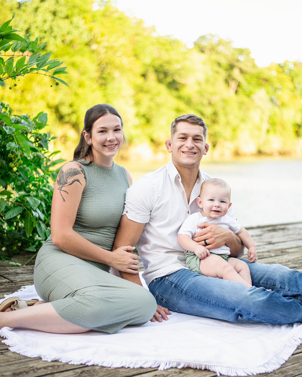 Young family with baby sits on white blanket on lakeside dock.