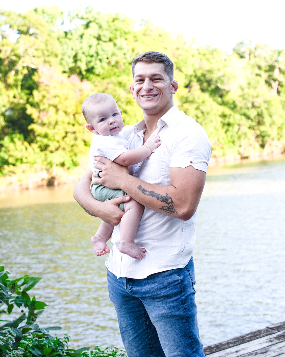 Smiling father holds happy baby boy on sunny lakeside wooden dock.
