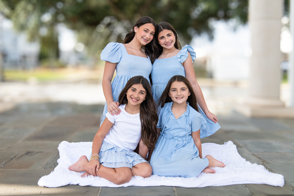 Four young girls in matching blue dresses pose together outdoors on white blanket for family portrait.
