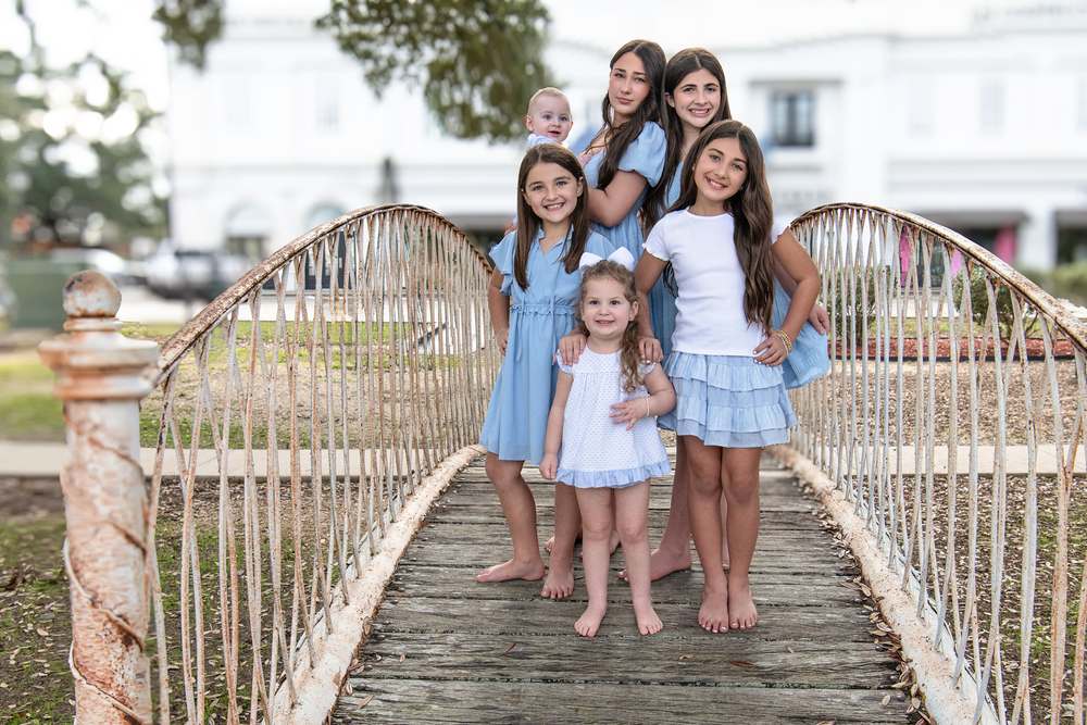 Six girls in blue and white outfits pose together on a charming wooden bridge outdoors.