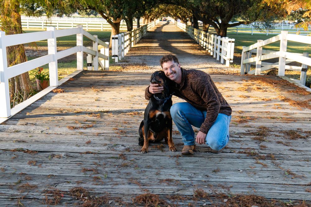 Man crouching with a Rottweiler dog on a wooden bridge path lined with white fences.