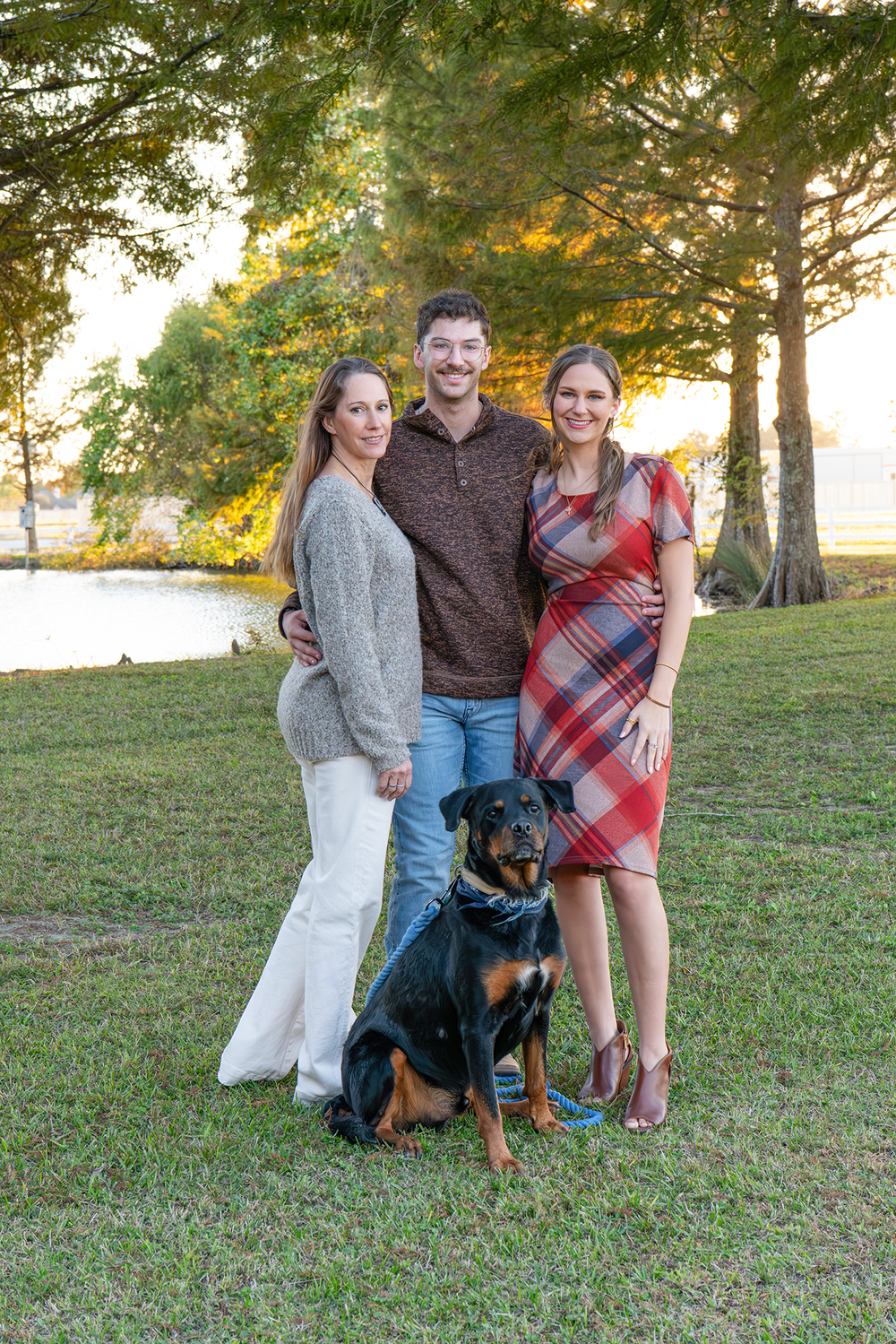 Three adults and Rottweiler dog pose together at golden hour lakeside.