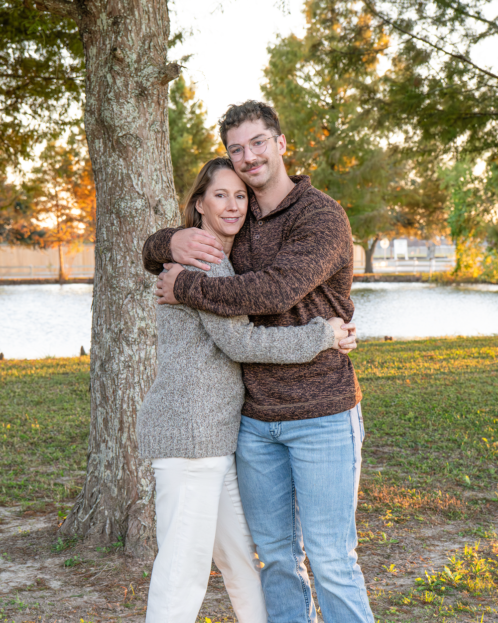 Couple hugging by a tree near a lake at sunset, man in brown sweater and woman in gray.