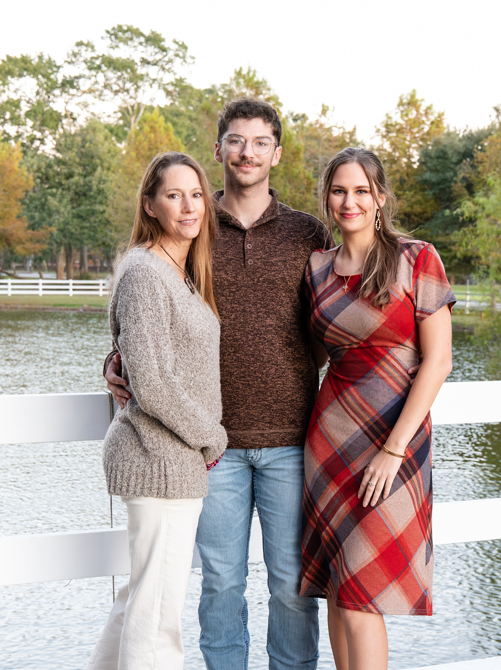Three adults posing together by a white fence near a lake during golden hour in autumn.