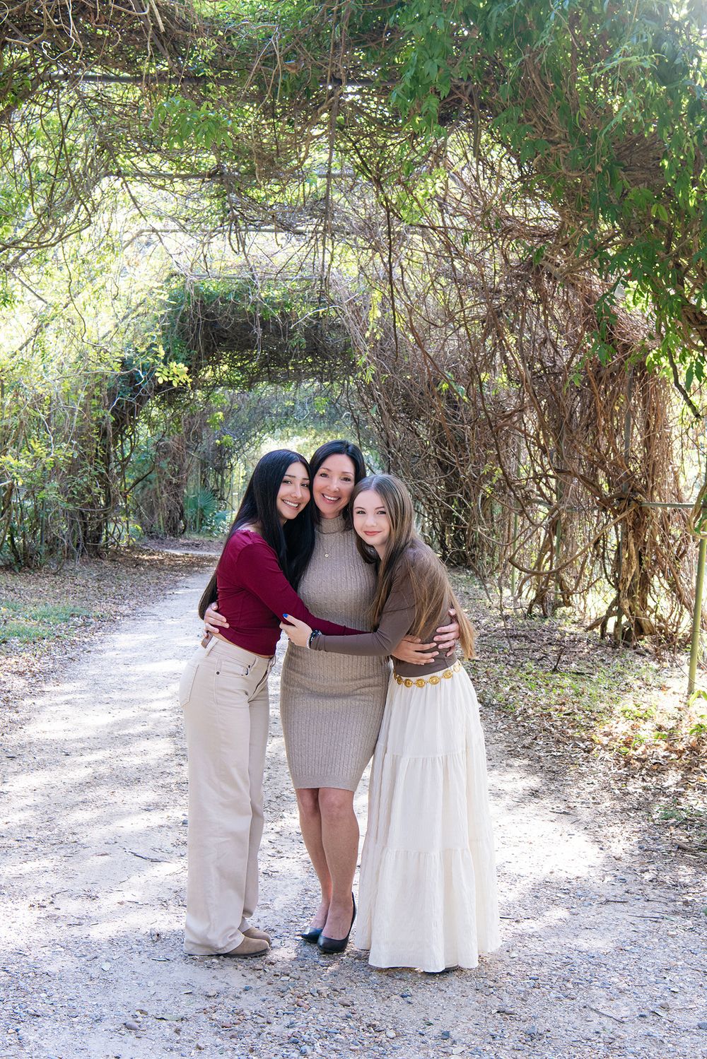 Mother embraces two teenage daughters under lush green garden archway.