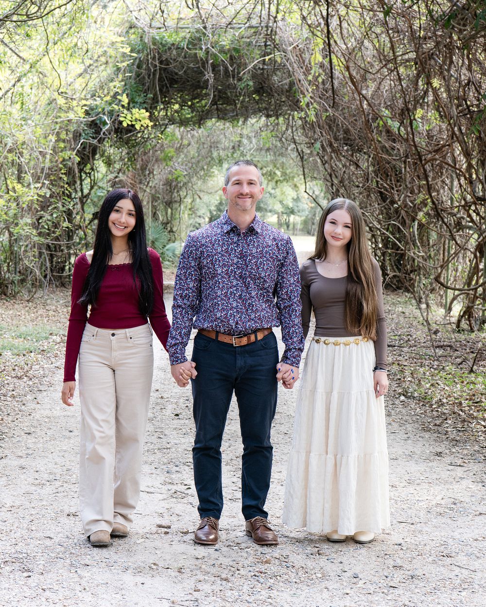 Father holds hands with two daughters on sunlit garden pathway.