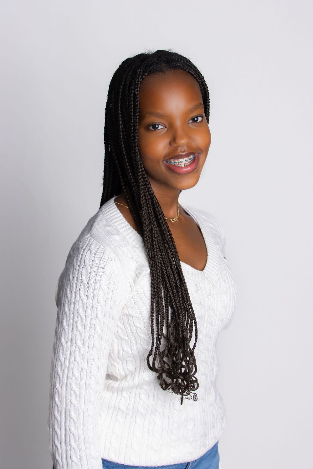 Person with long braided hairstyle wearing a white cable knit sweater poses against a gray background.