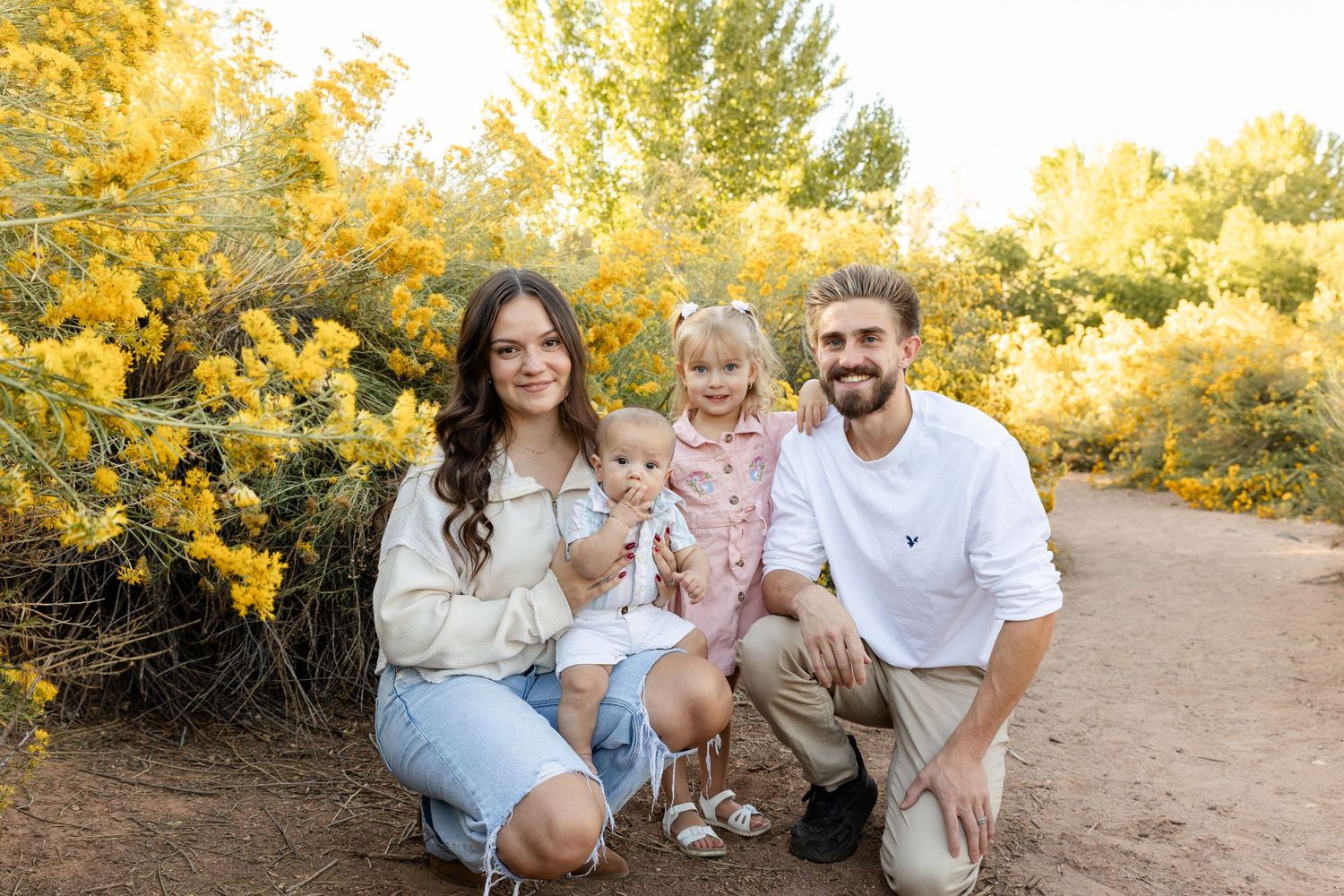 Smiling family of four posing together among bright yellow flowering shrubs in golden afternoon light.