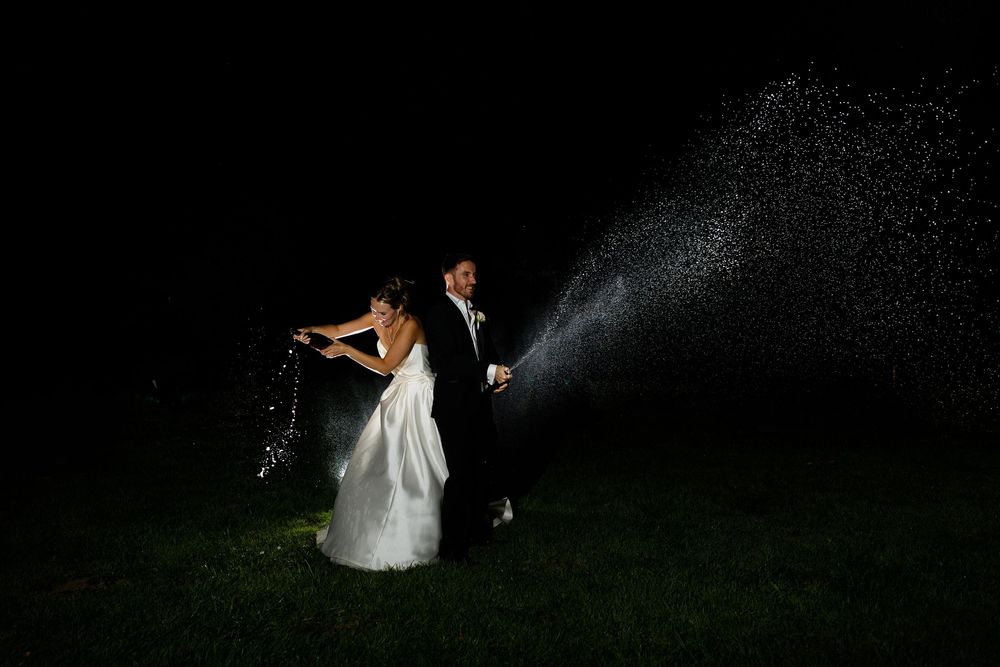 A couple in wedding attire stands in darkness while being illuminated by spraying water on a grassy lawn.