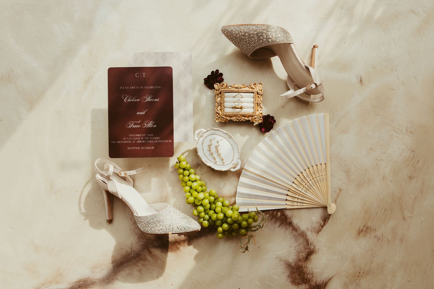 Bridal flat lay featuring lace heels, wedding invitation, hand fan, ring, and green grapes on marble.