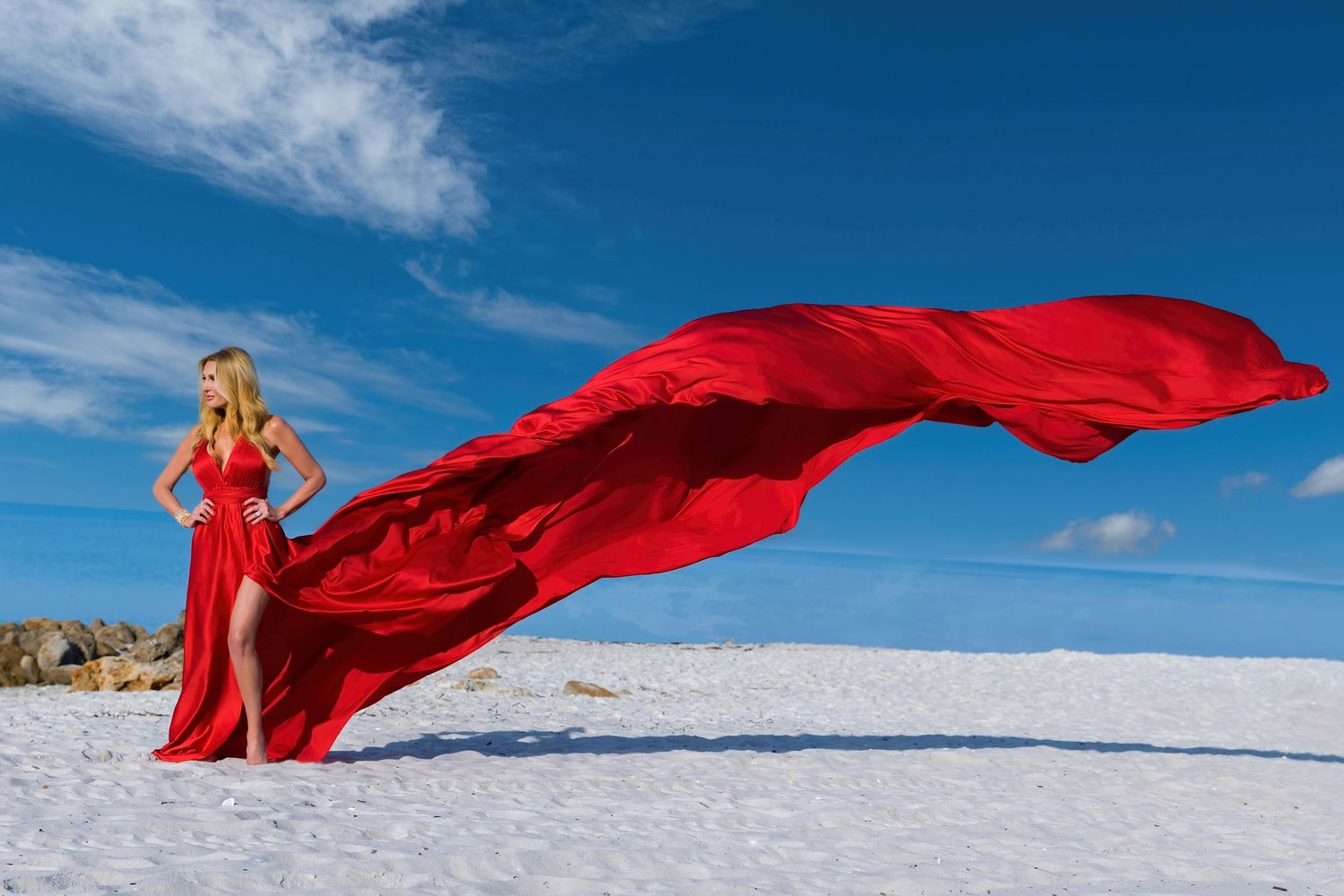A flowing red dress billows dramatically against a snowy winter landscape beneath a bright blue sky.