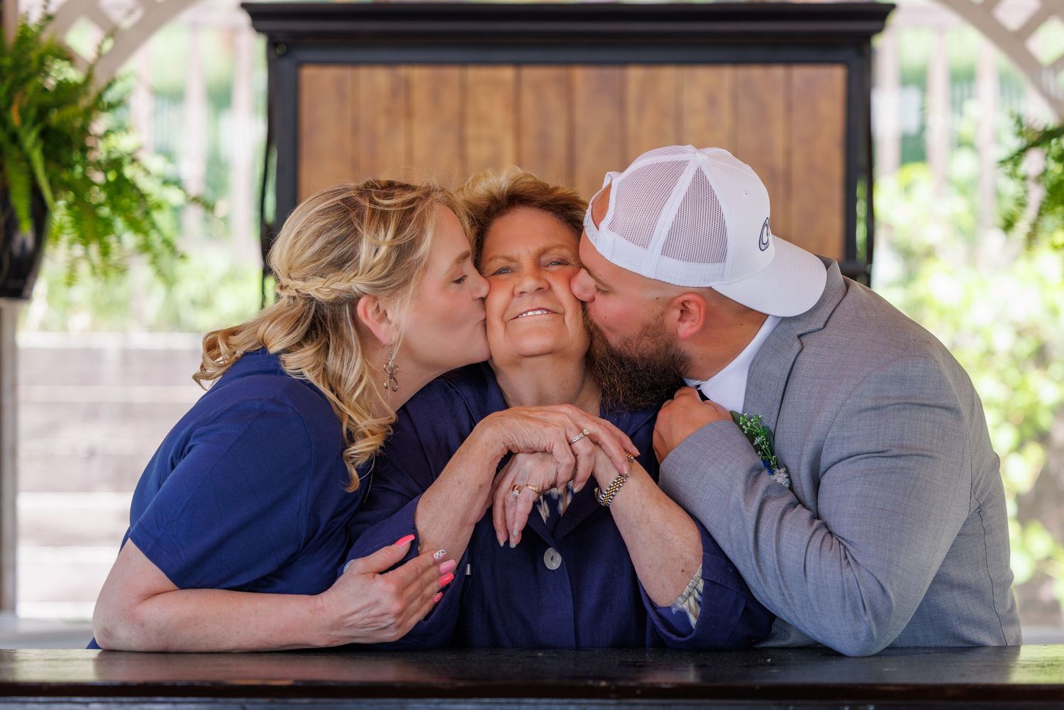 Three people in casual clothing share an affectionate moment together on a porch with greenery in the background.