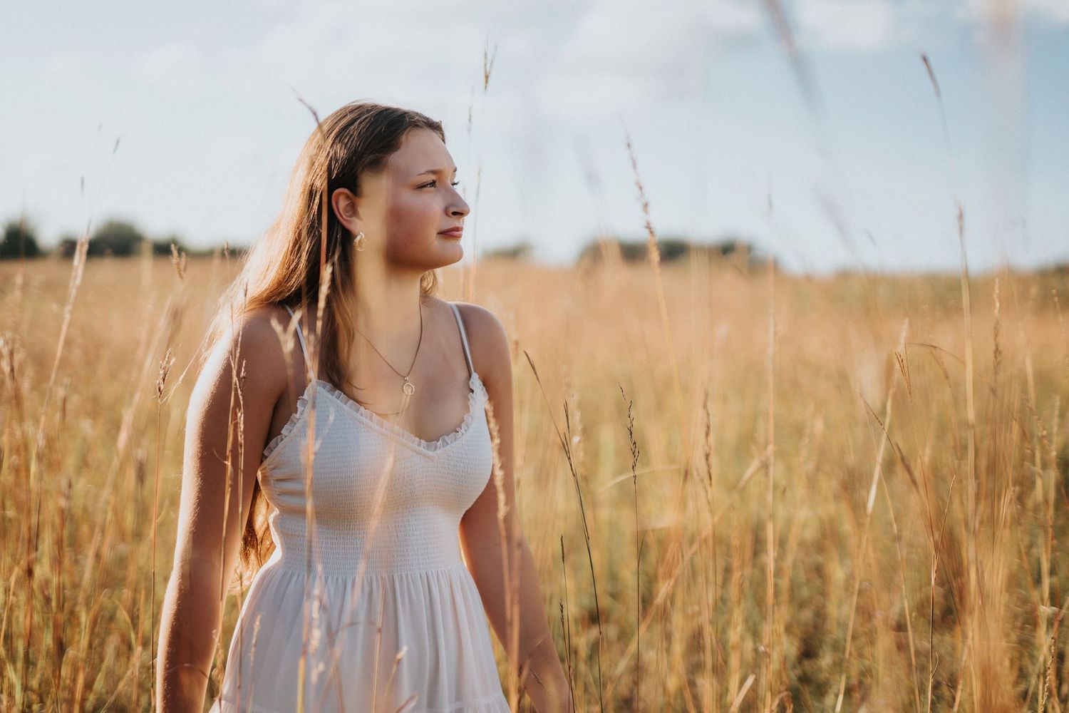 A woman in a white dress stands in a golden wheat field at sunset with warm sunlight illuminating the scene.