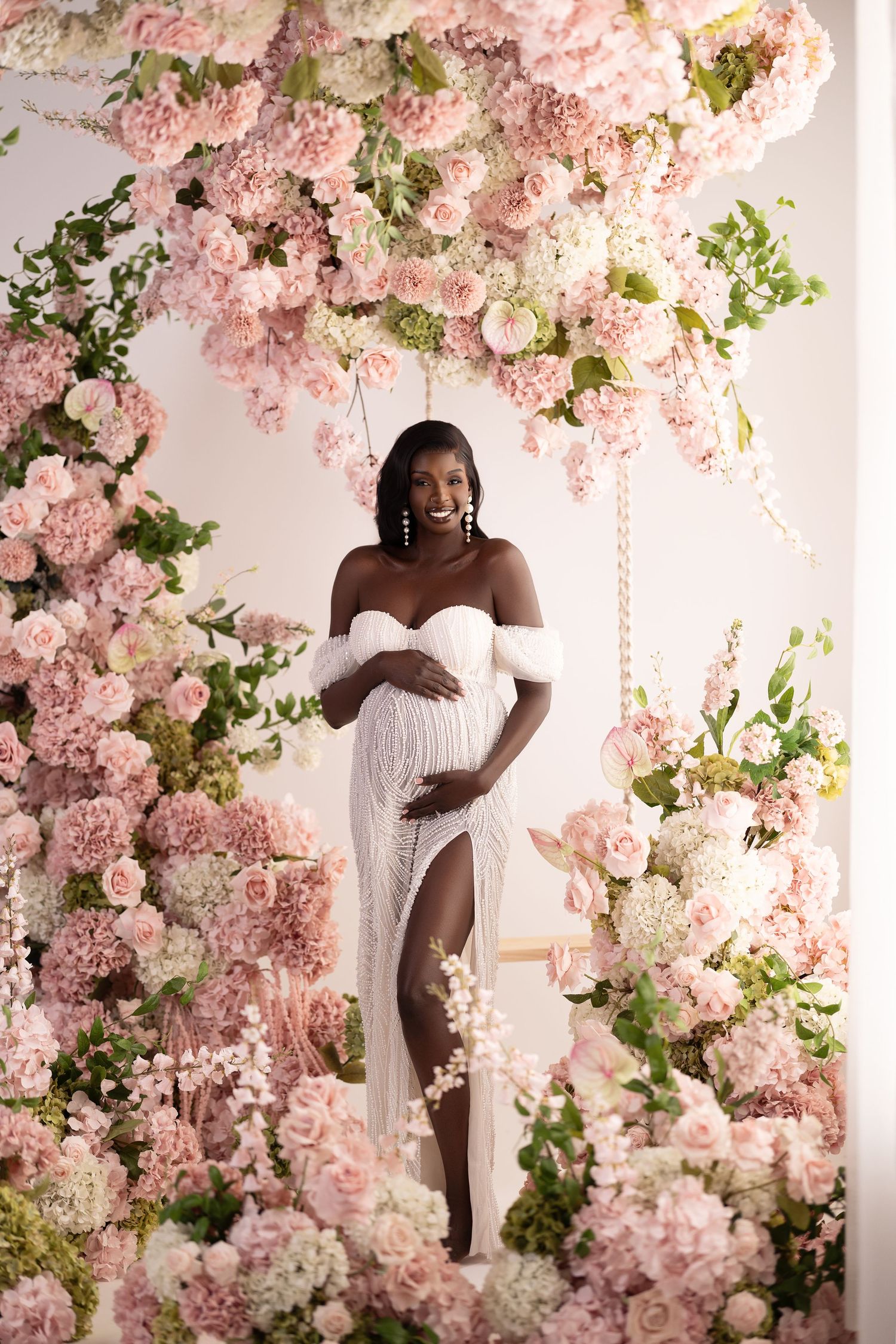 Elegant maternity photo with flowing white dress against a romantic floral arch of pink and white blossoms.