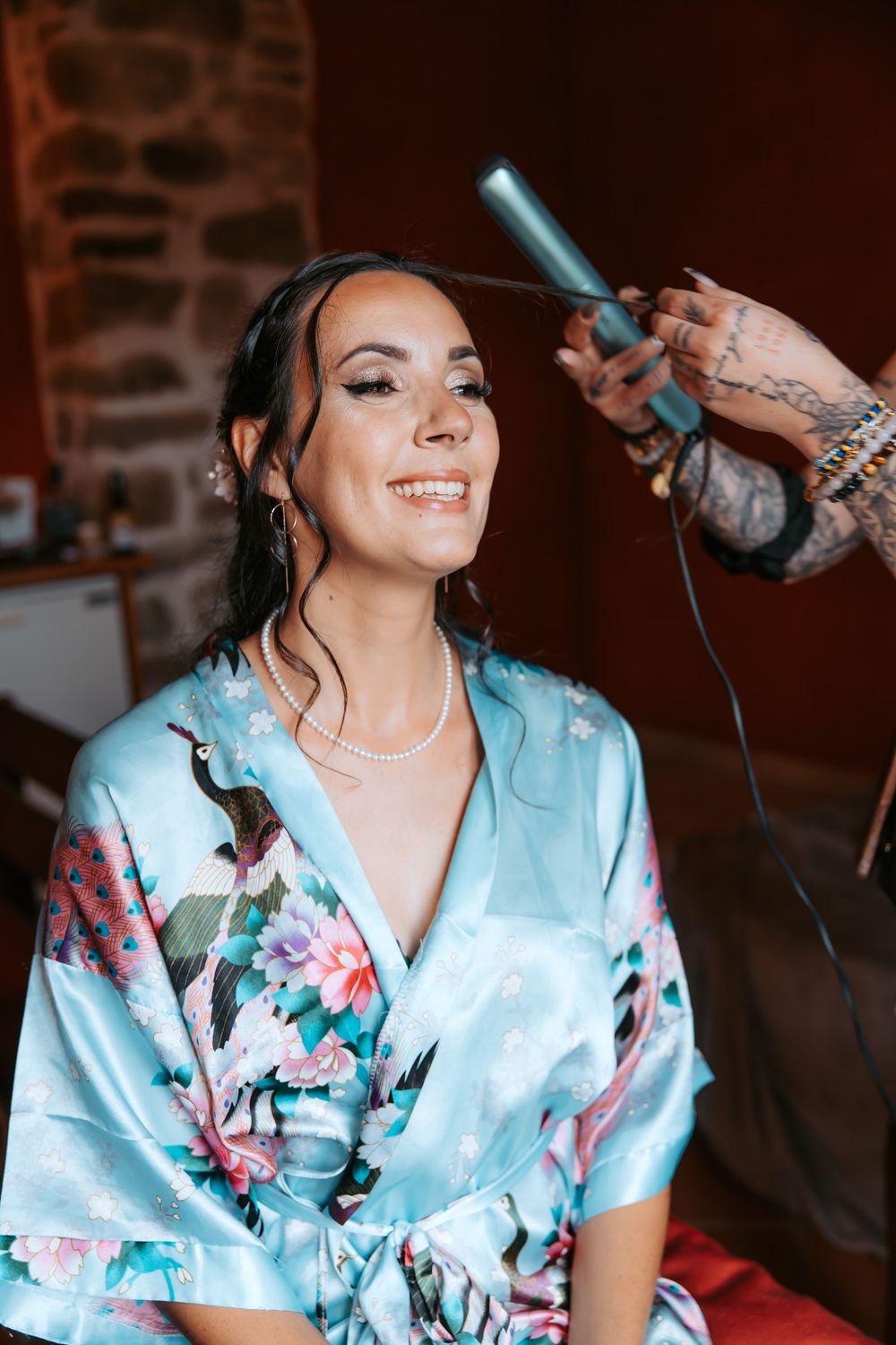 A smiling person in a floral silk robe gets their hair styled while wearing a necklace during wedding preparations.