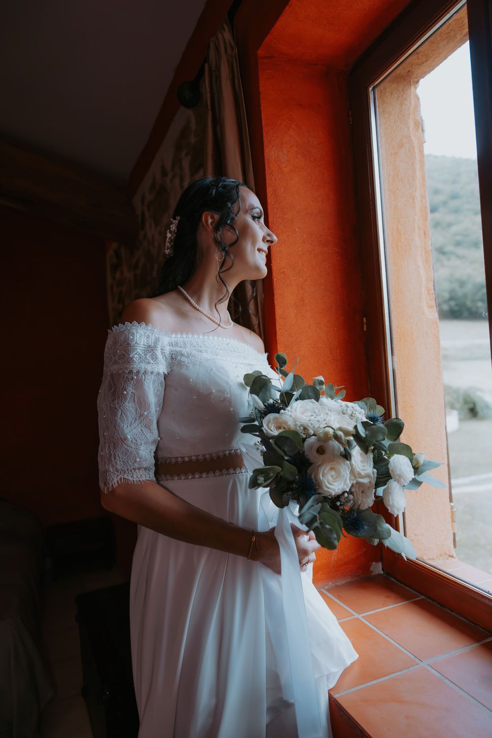 Bride in white lace gown holds eucalyptus bouquet while gazing out window in warm orange-lit room.