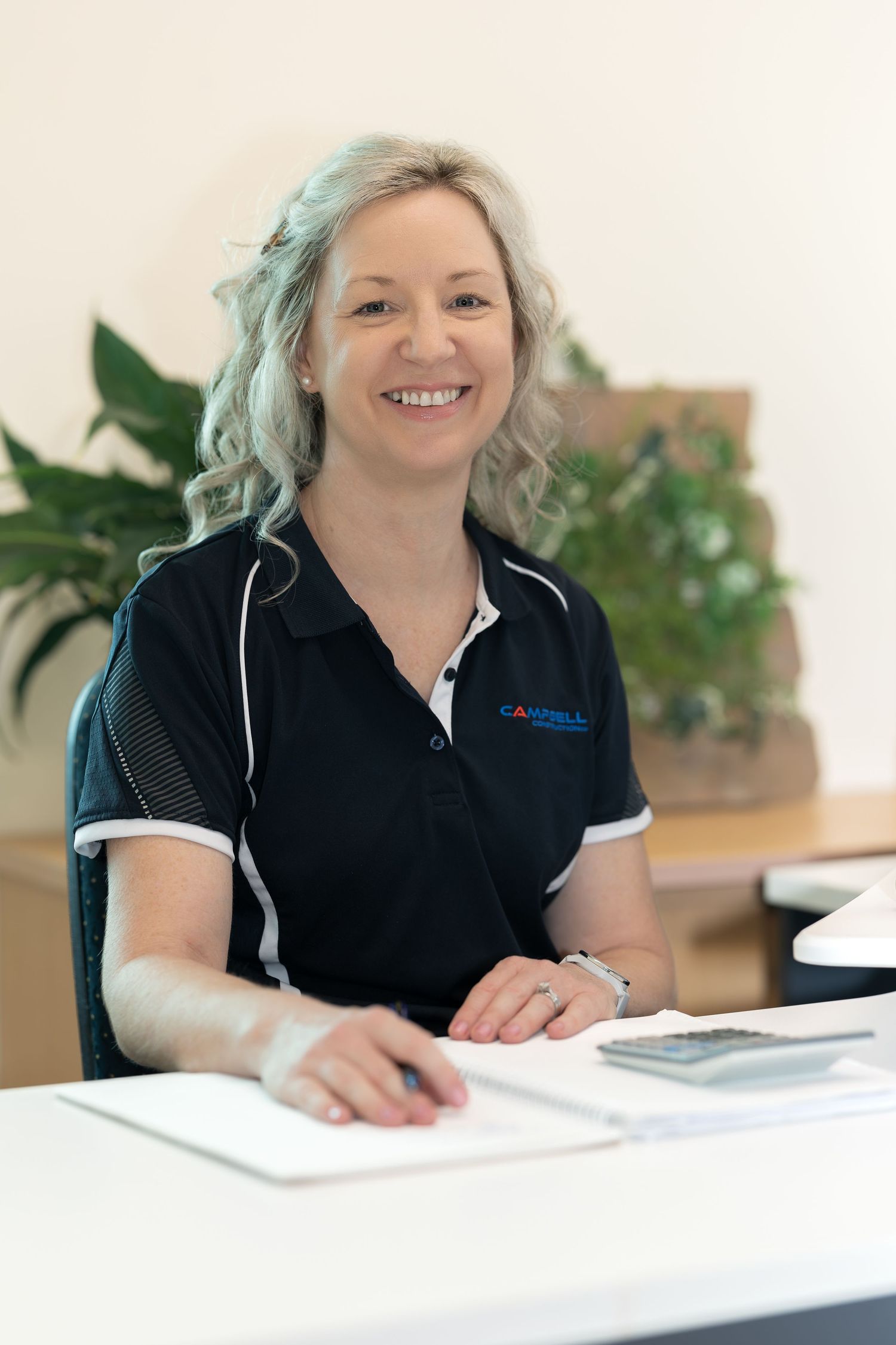 Professional staff member in a navy uniform polo shirt smiles while working at a white desk with papers and office plants nearby.