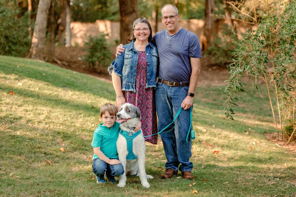 Family and pet dog posing together for casual outdoor portraits on grassy lawn with trees in background.