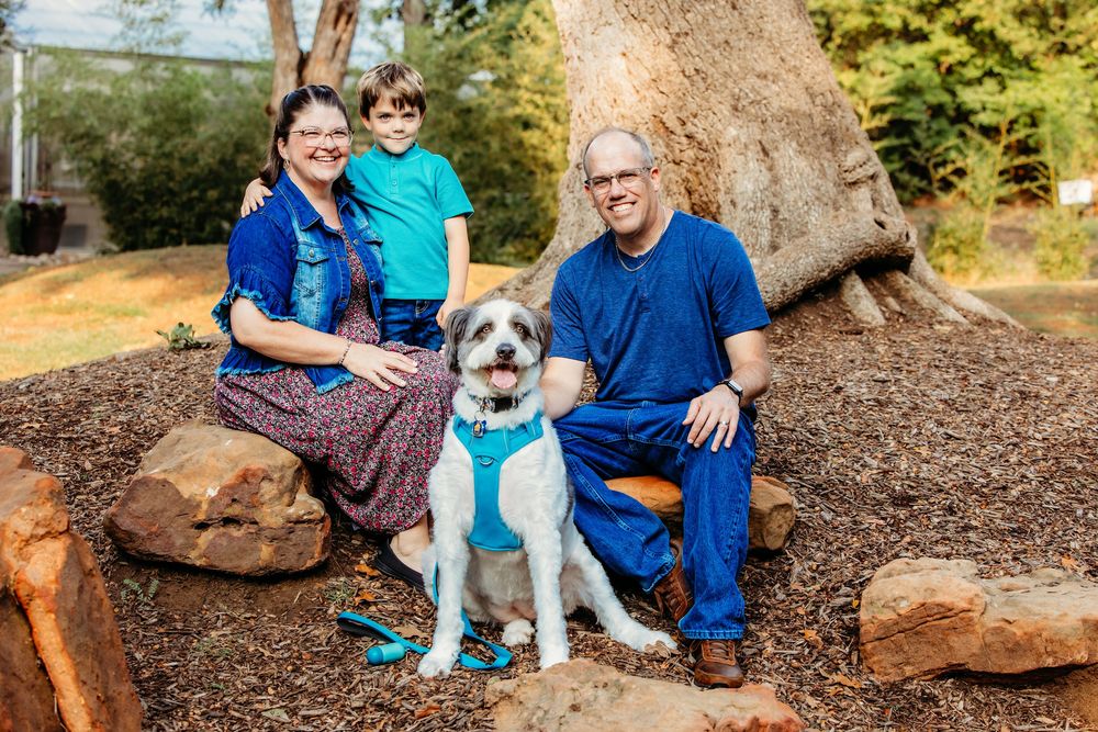 A family and their white dog pose together on a tree-lined path in coordinating blue outfits during an outdoor photo session.