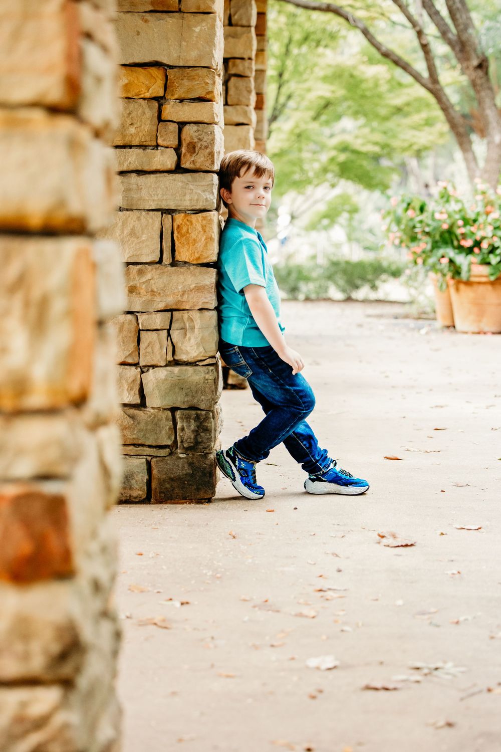 A child in a turquoise shirt and blue sneakers leans against a stone pillar on a garden path.