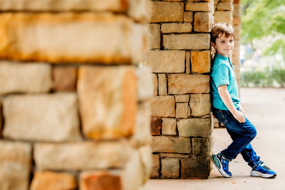 Person in turquoise shirt and jeans sits on stone wall while looking down at their feet in casual outdoor setting.