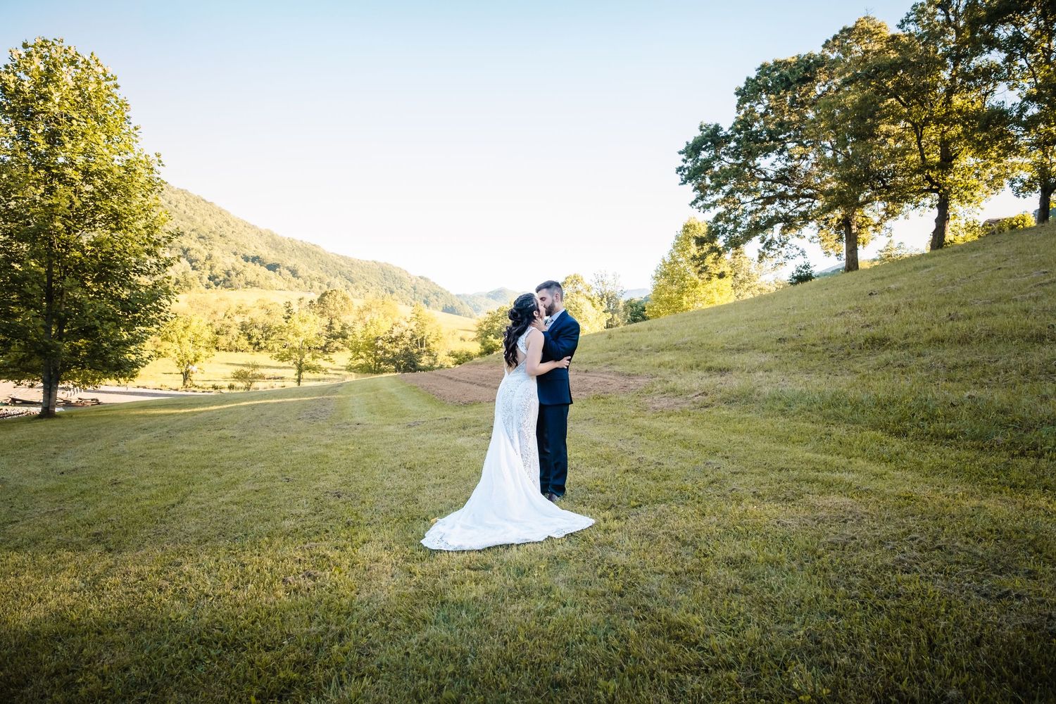 A couple in wedding attire embraces on a grassy hillside during golden hour at an outdoor venue.