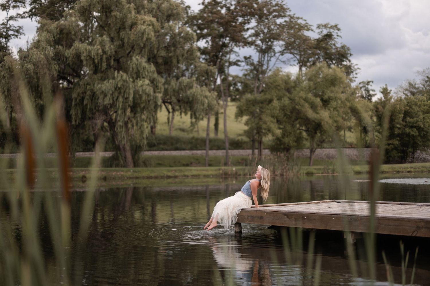 A wooden boardwalk extends over a tranquil lake surrounded by willow trees and natural vegetation.