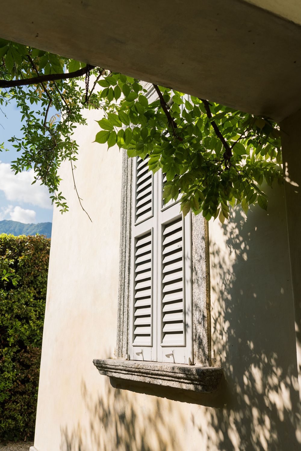 White shuttered windows on a villa exterior with dappled sunlight filtering through leaves.