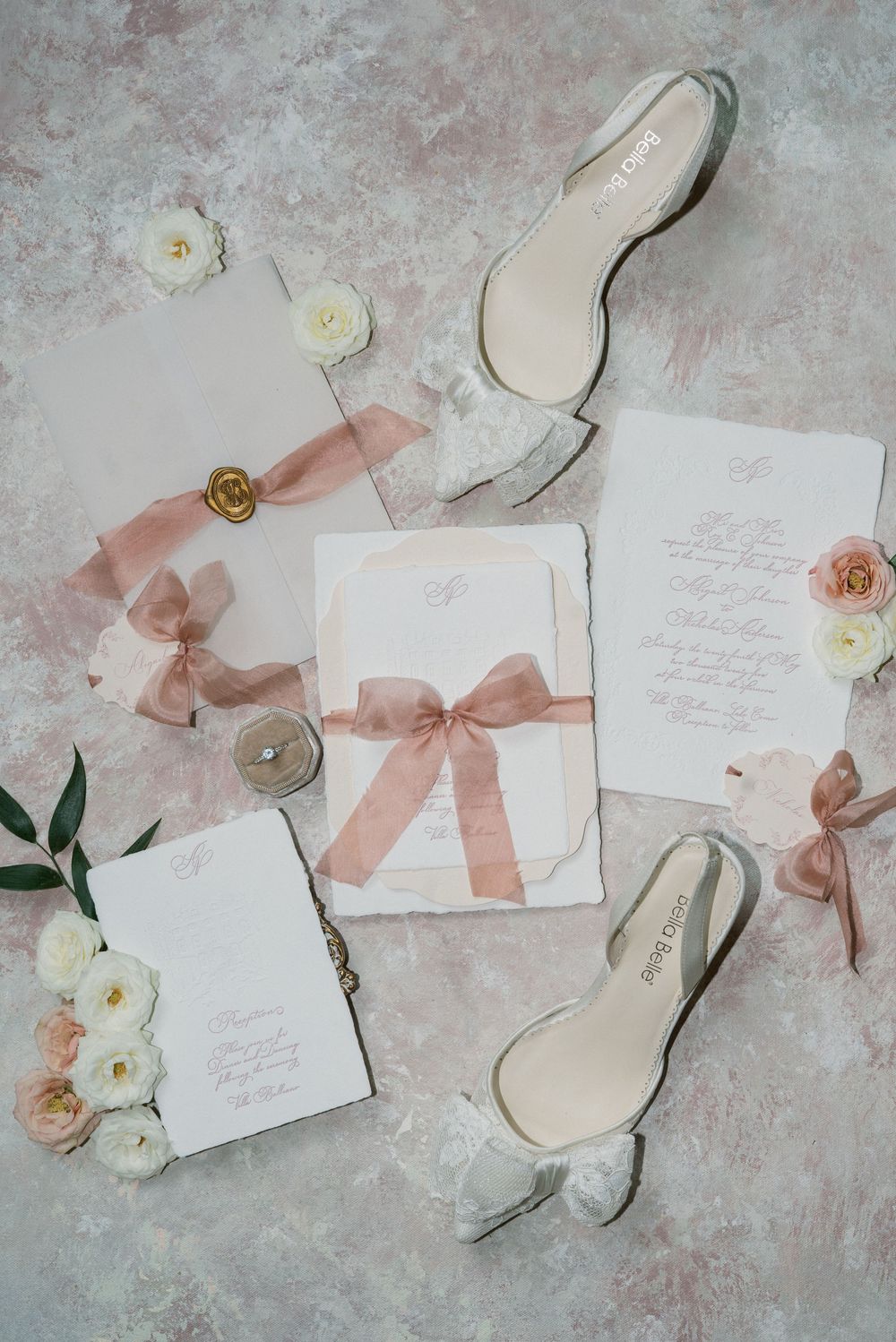 Wedding invitations with blush pink ribbons and white roses on a textured gray background.