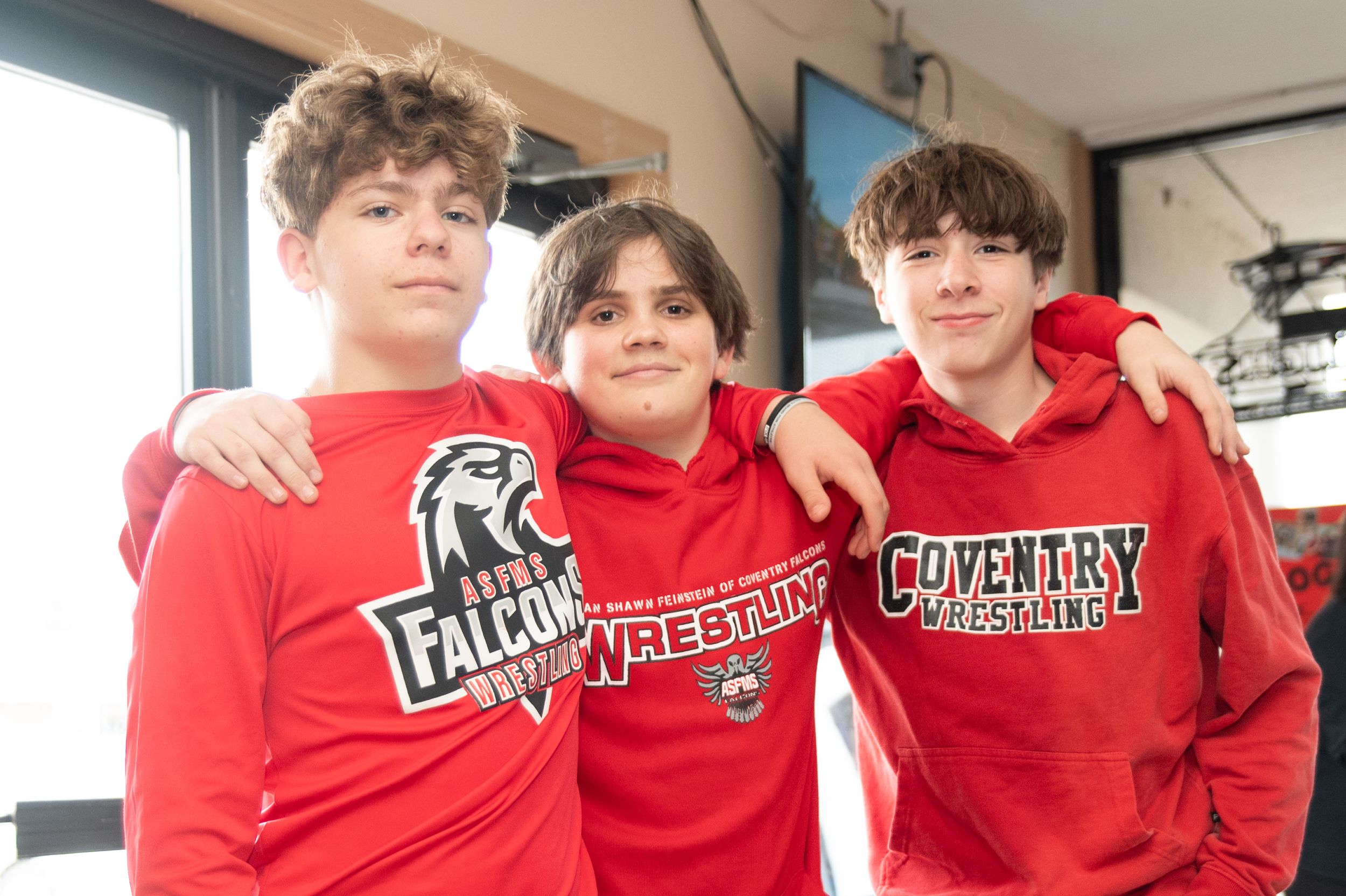 Three students wearing matching red Falconry hooded sweatshirts pose together indoors.