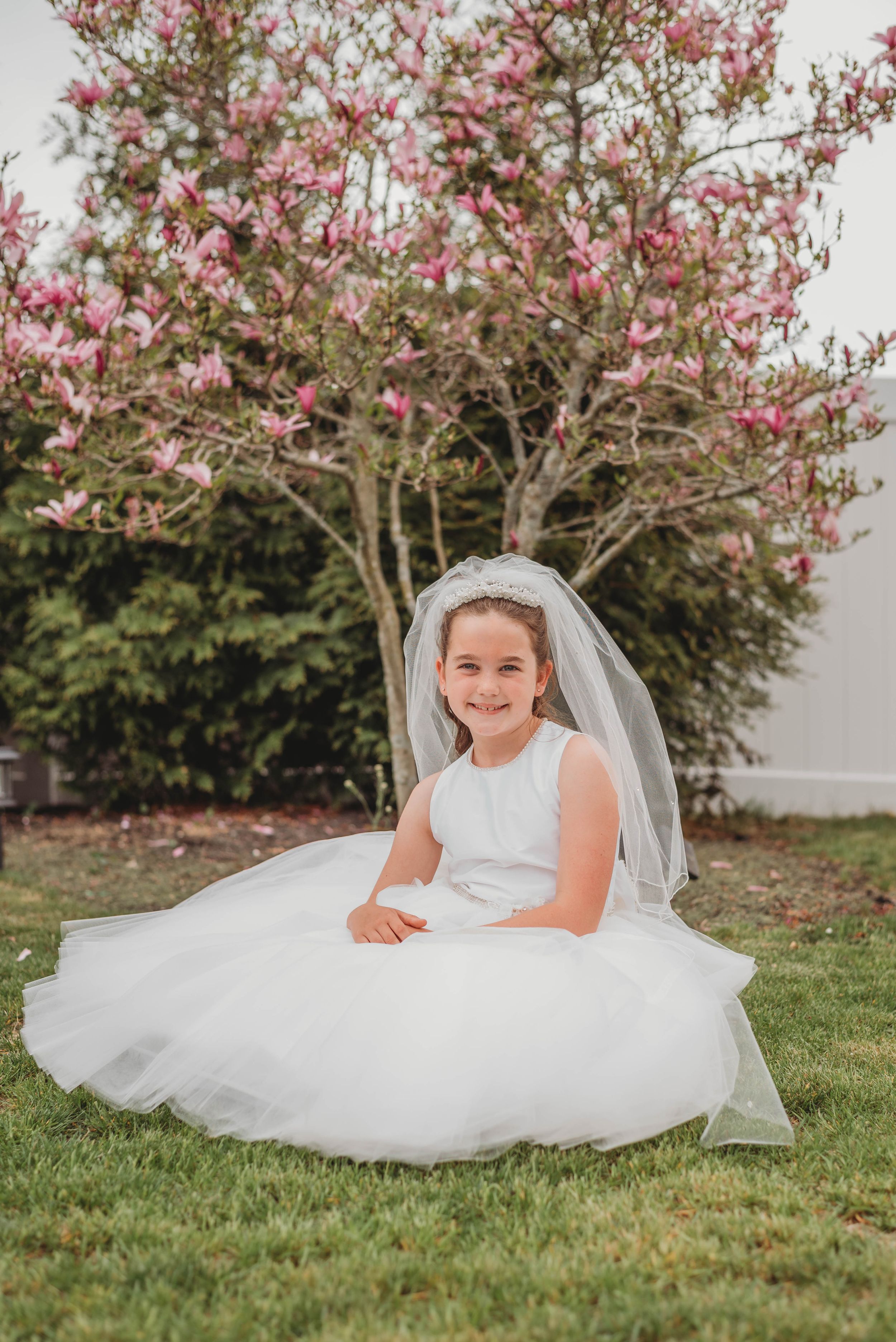 A young figure in a white dress and veil poses gracefully on grass beneath blooming pink magnolia trees.