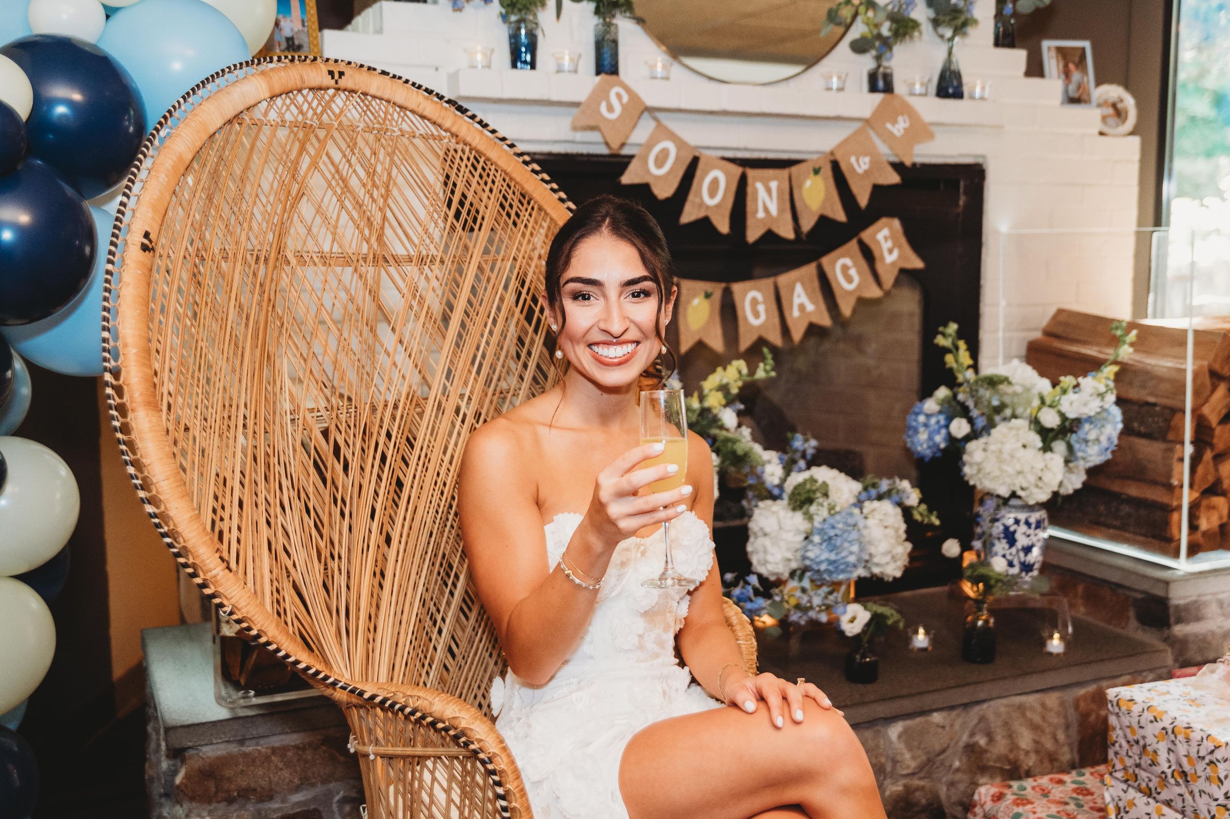 Bride sits in rattan peacock chair surrounded by wedding decor and balloons at bridal shower celebration.