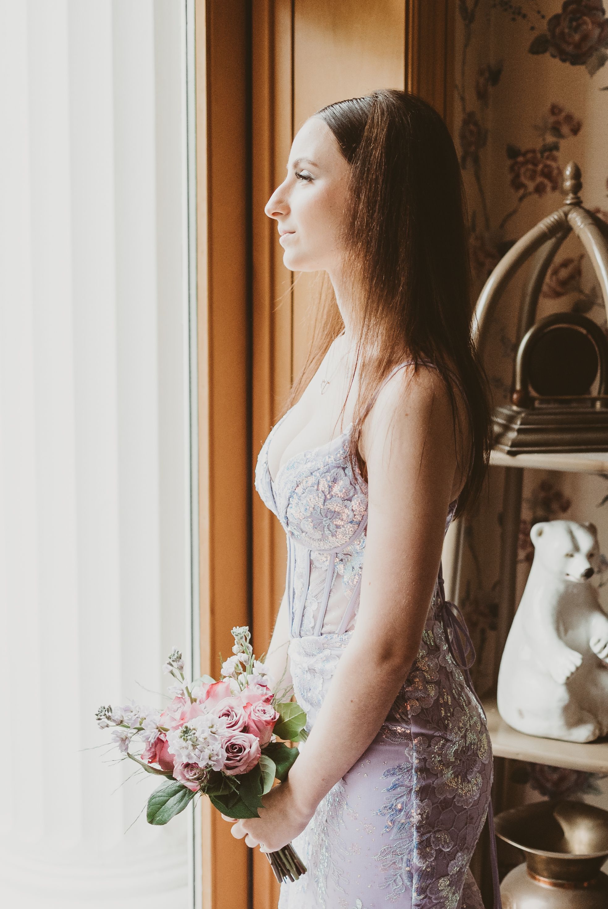 A bride in a fitted lace wedding dress stands by a window holding a pink and white bouquet in natural light.