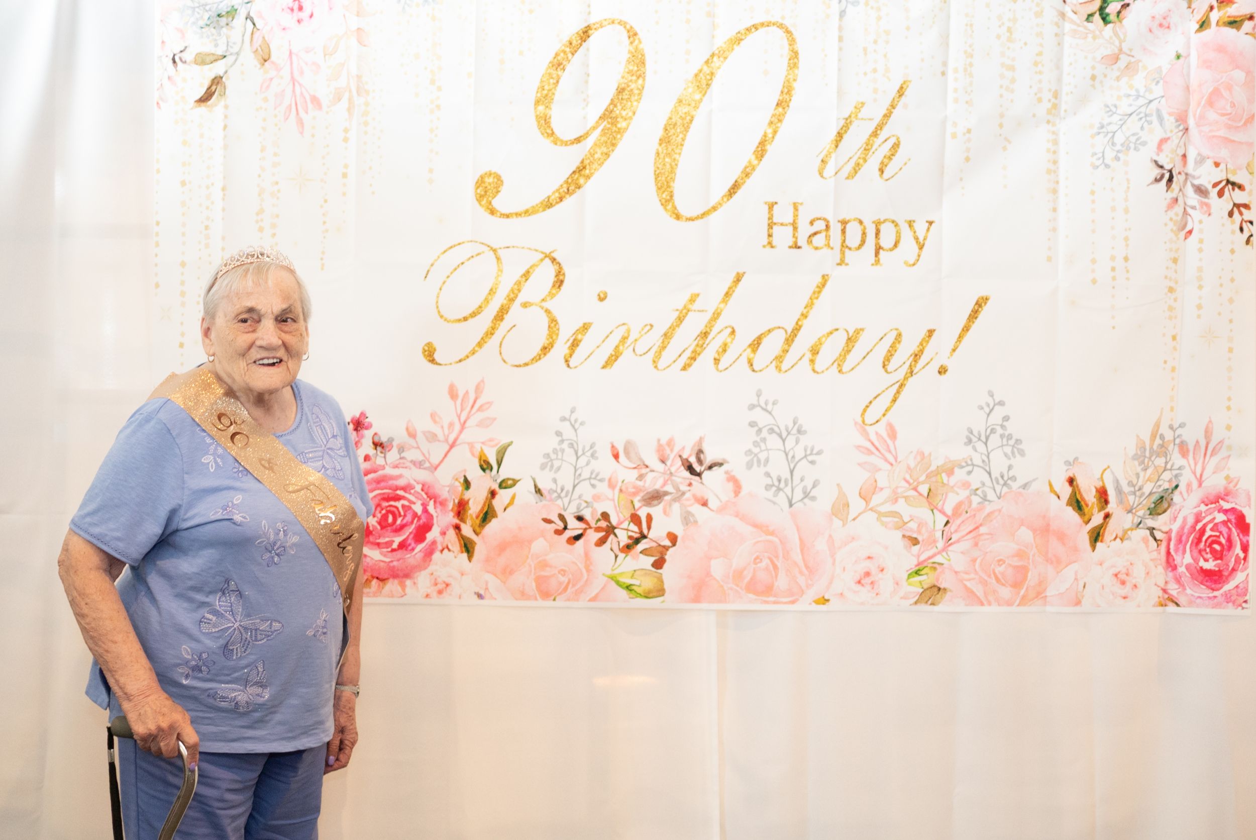 A 90th birthday celebration backdrop with pink roses and gold lettering decorates an elegant party setting.