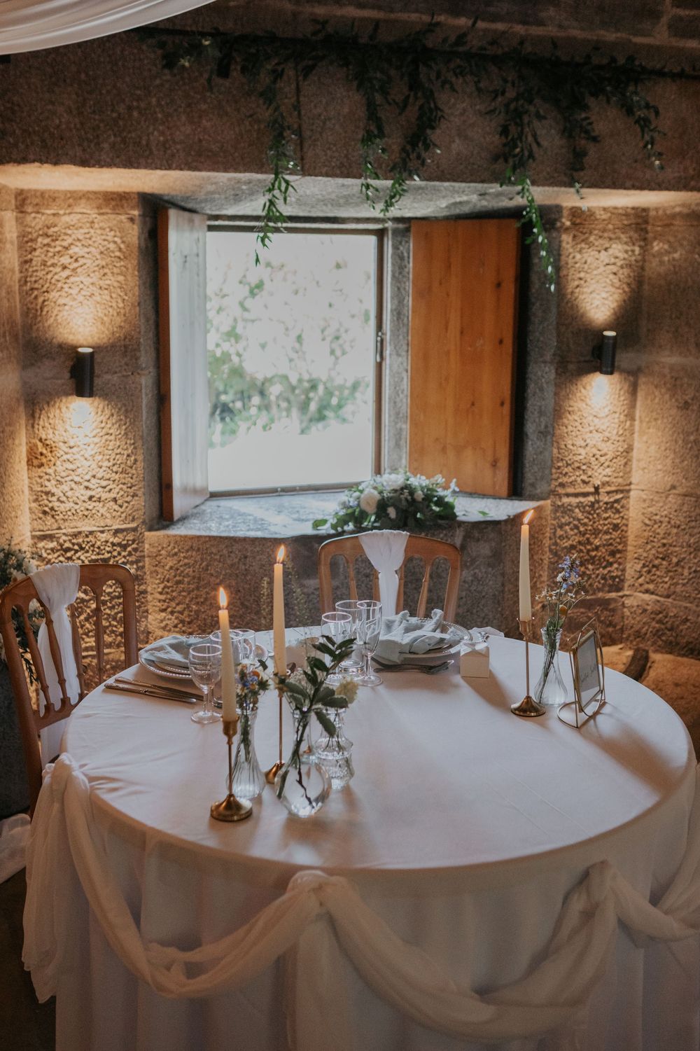 Elegant wedding sweetheart table with white linens, candles, and greenery in stone venue.