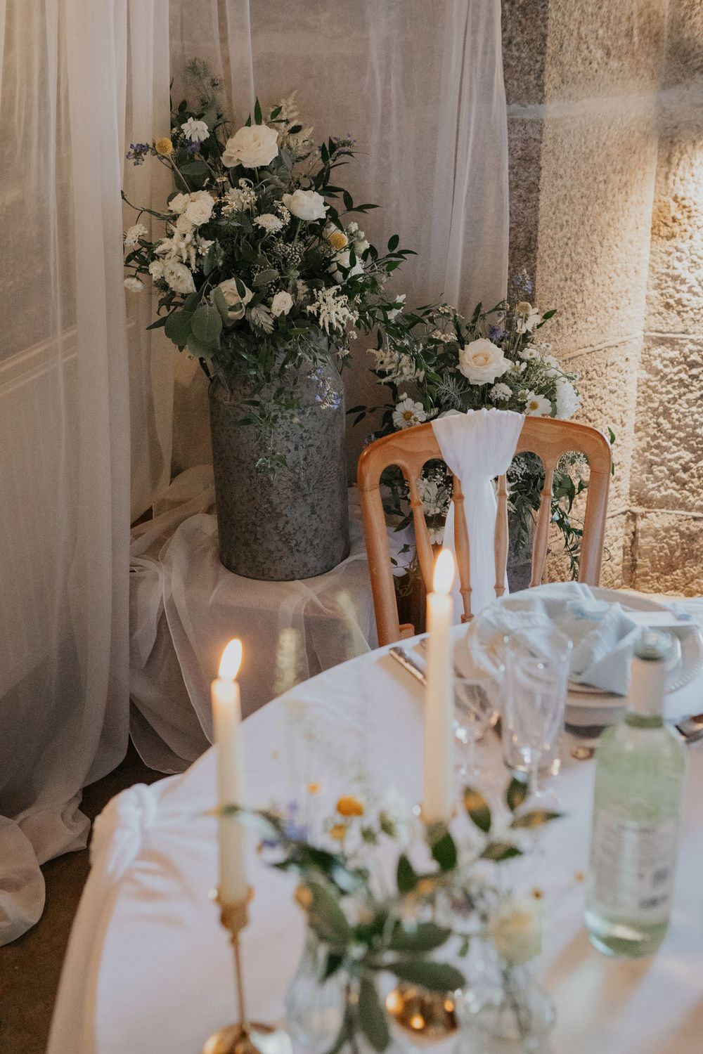 Wedding decor featuring large white floral arrangement in vintage vase beside candlelit table.