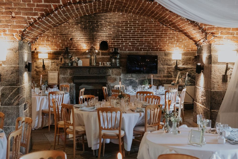 Rustic wedding reception in a brick vaulted venue with round tables, white linens, and a stone fireplace.