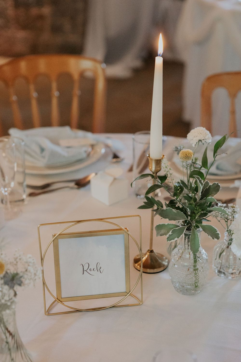 Elegant wedding table setting with gold geometric frame table number, white taper candle, and floral arrangement.