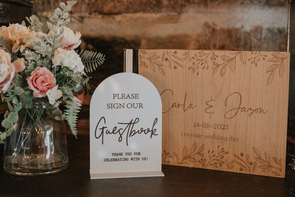 Wedding guestbook sign beside a wooden engraved keepsake board for Carla and Jason dated 14-08-2023.