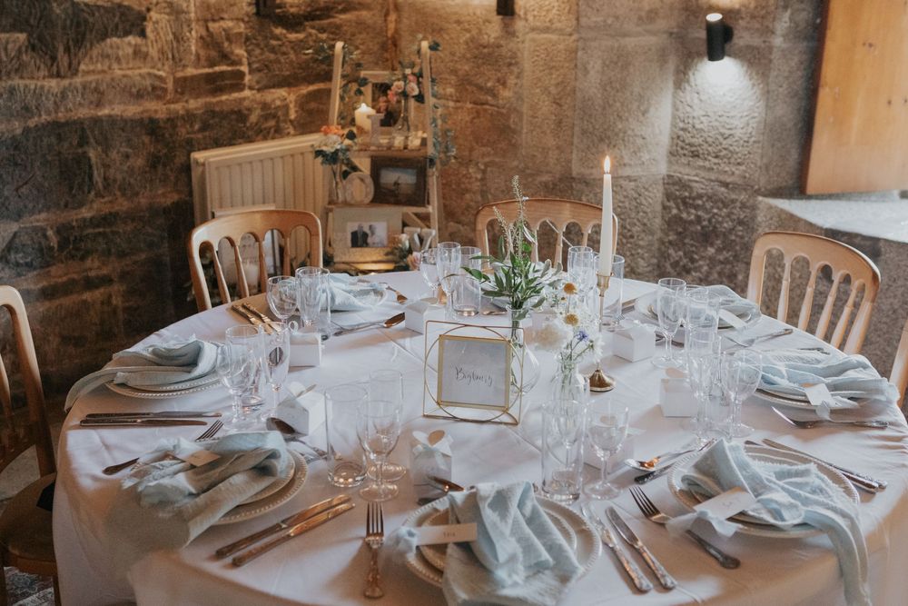 Round wedding reception table with white linens, sage napkins, candles, and a gold table number frame.