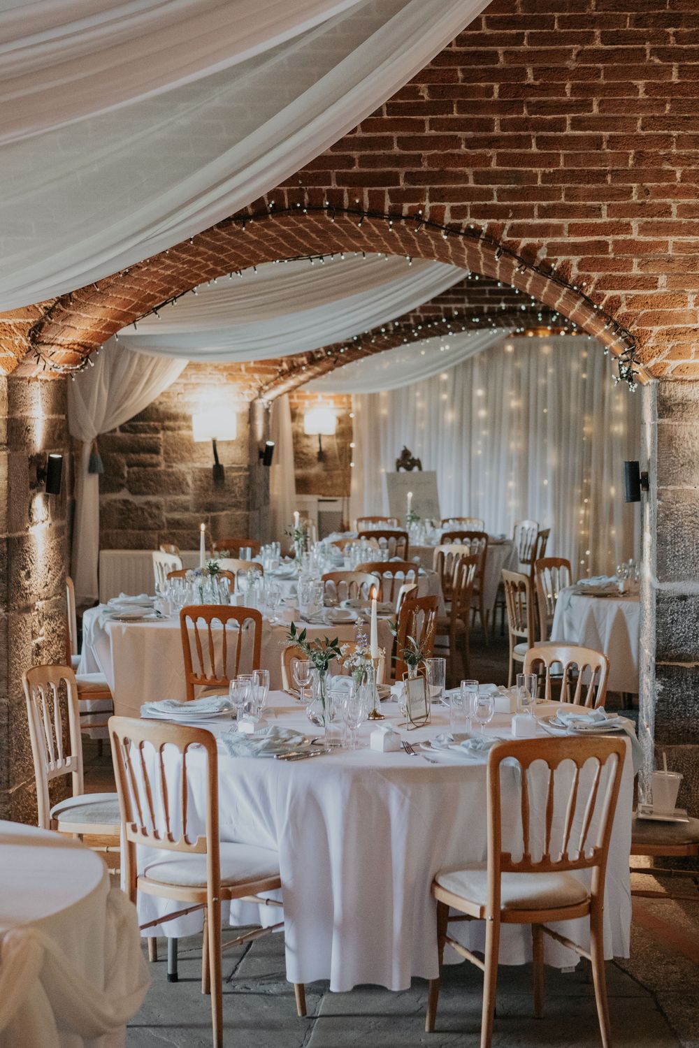 Romantic wedding venue with brick arches, fairy lights, white draping, and elegantly set round tables.