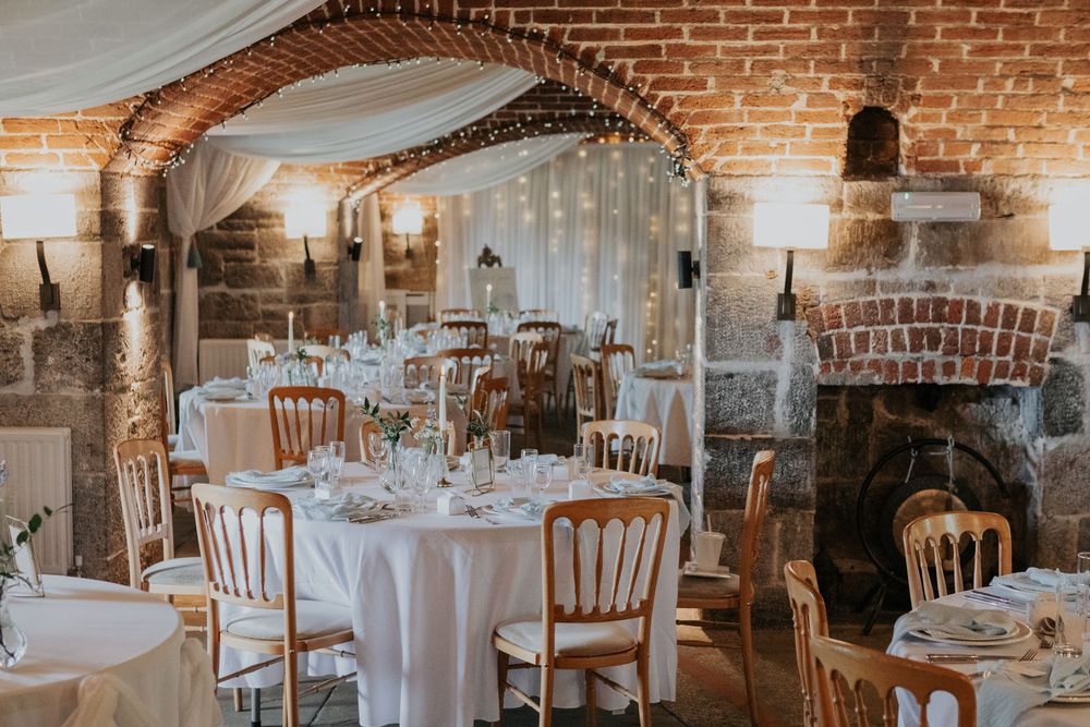 Wedding reception hall with exposed brick walls, arched doorways, fairy lights, and white-clothed round tables.