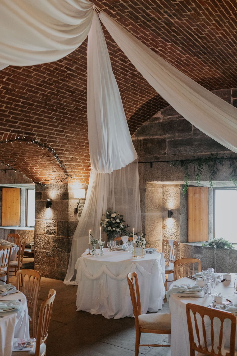 Wedding venue interior with vaulted brick ceiling, flowing white ceiling drapes, and elegantly set tables.