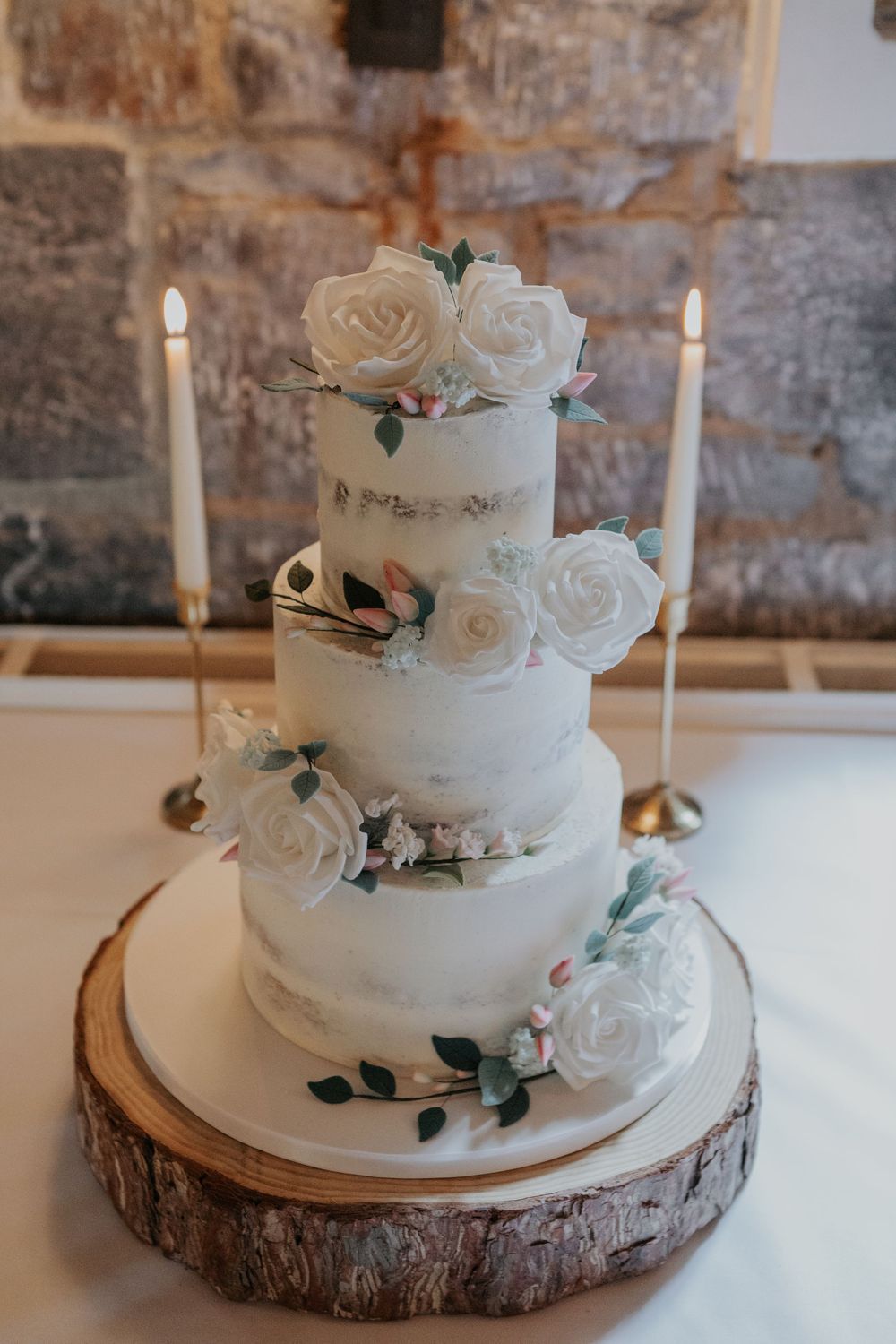 Three-tier semi-naked wedding cake decorated with white sugar roses on a rustic wooden slice stand.