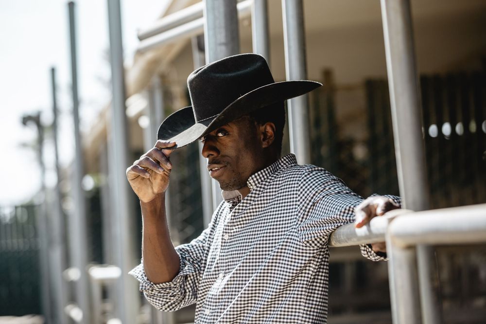 A person in a black cowboy hat and checkered shirt tips their hat while standing on a porch with palm trees in the background.