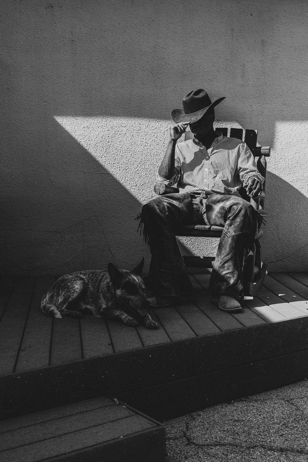A figure in a hat sits in sunlight on a wooden porch with a dog resting nearby in black and white photography.