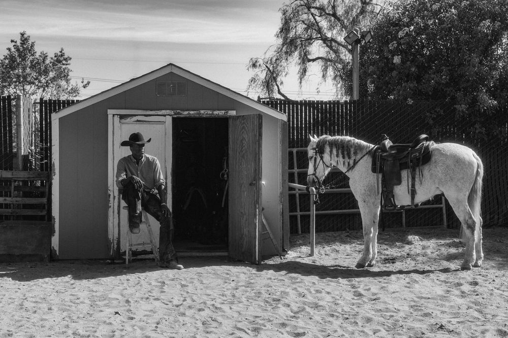White ranch buildings and stables line up against a desert landscape with horses visible nearby.
