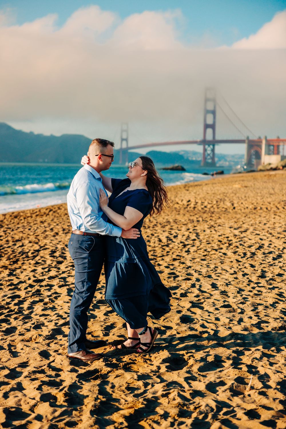 A romantic couple embraces on Baker Beach with the Golden Gate Bridge creating a stunning backdrop in San Francisco.