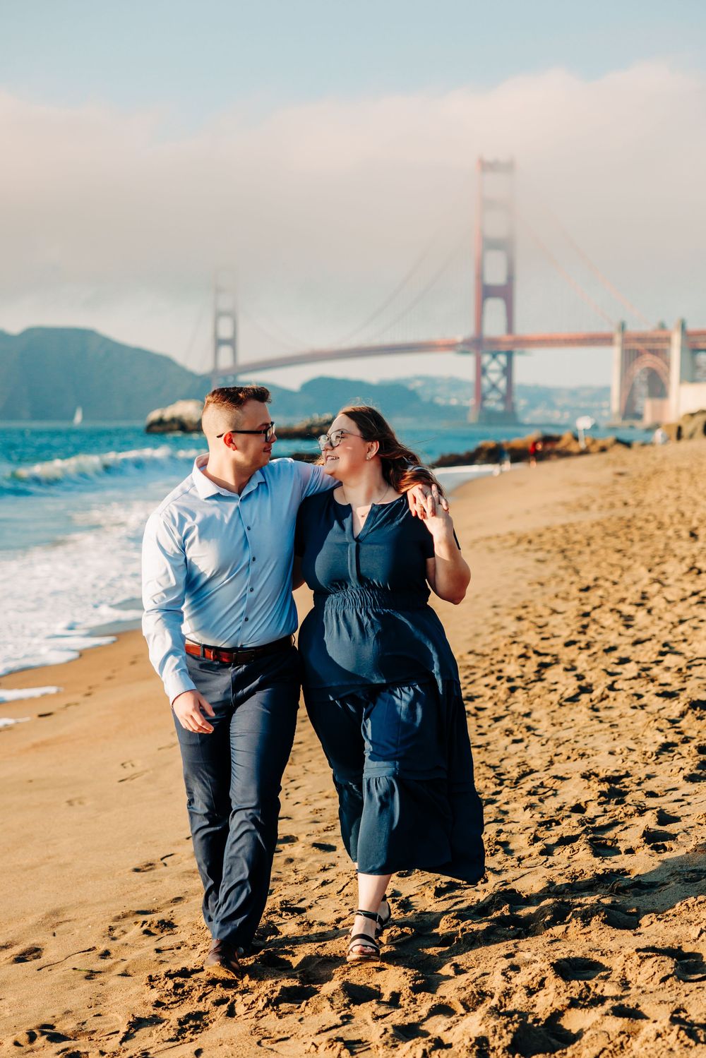 A couple strolls along Baker Beach's sandy shoreline with the iconic Golden Gate Bridge visible in the background.